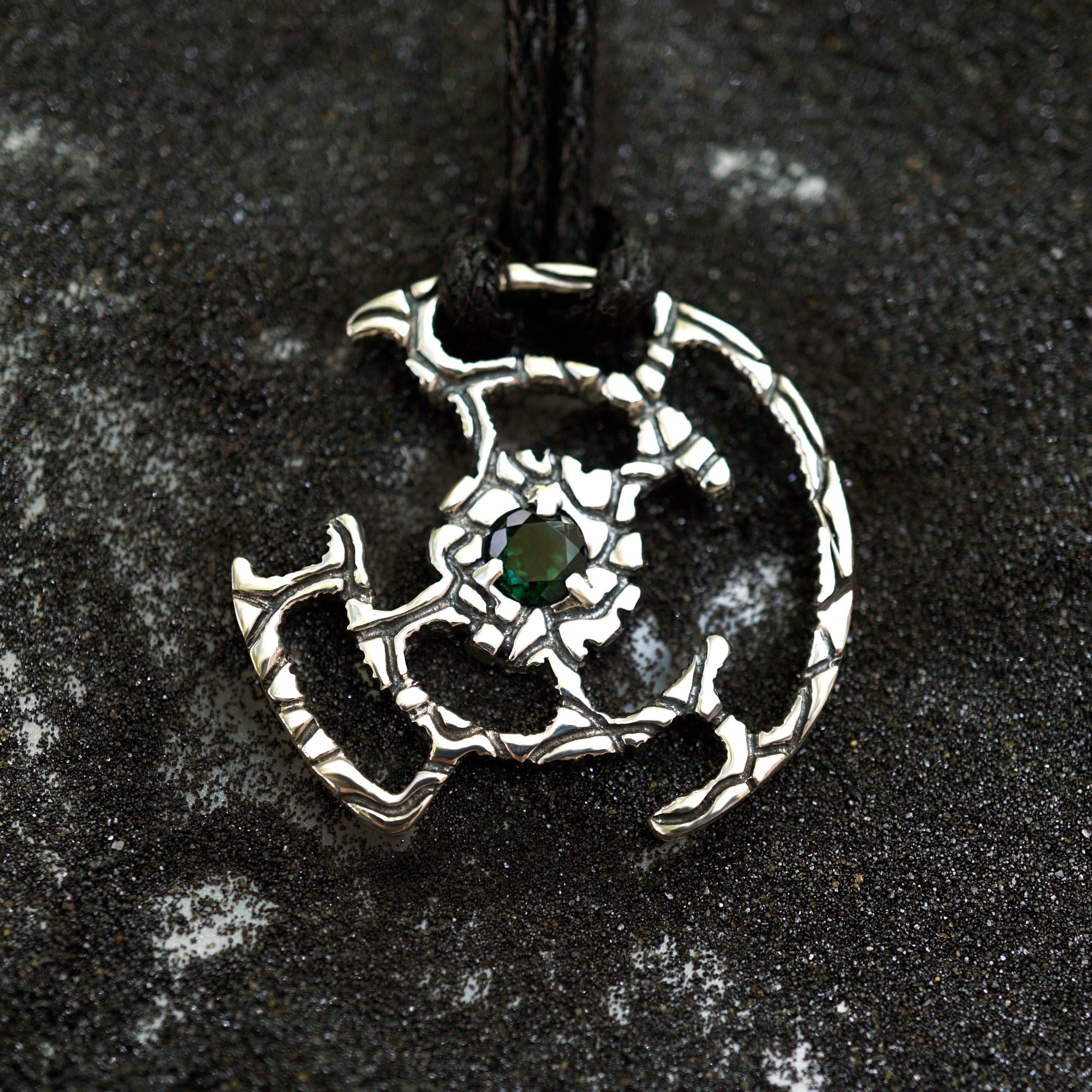 An oxidized solid sterling silver pendant with a green Tourmaline gemstone in the center. The pendant is in the shape of a circle with intricate designs. The pendant is hanging from a black cord. Cyberpunk design jewelry by Moonique.