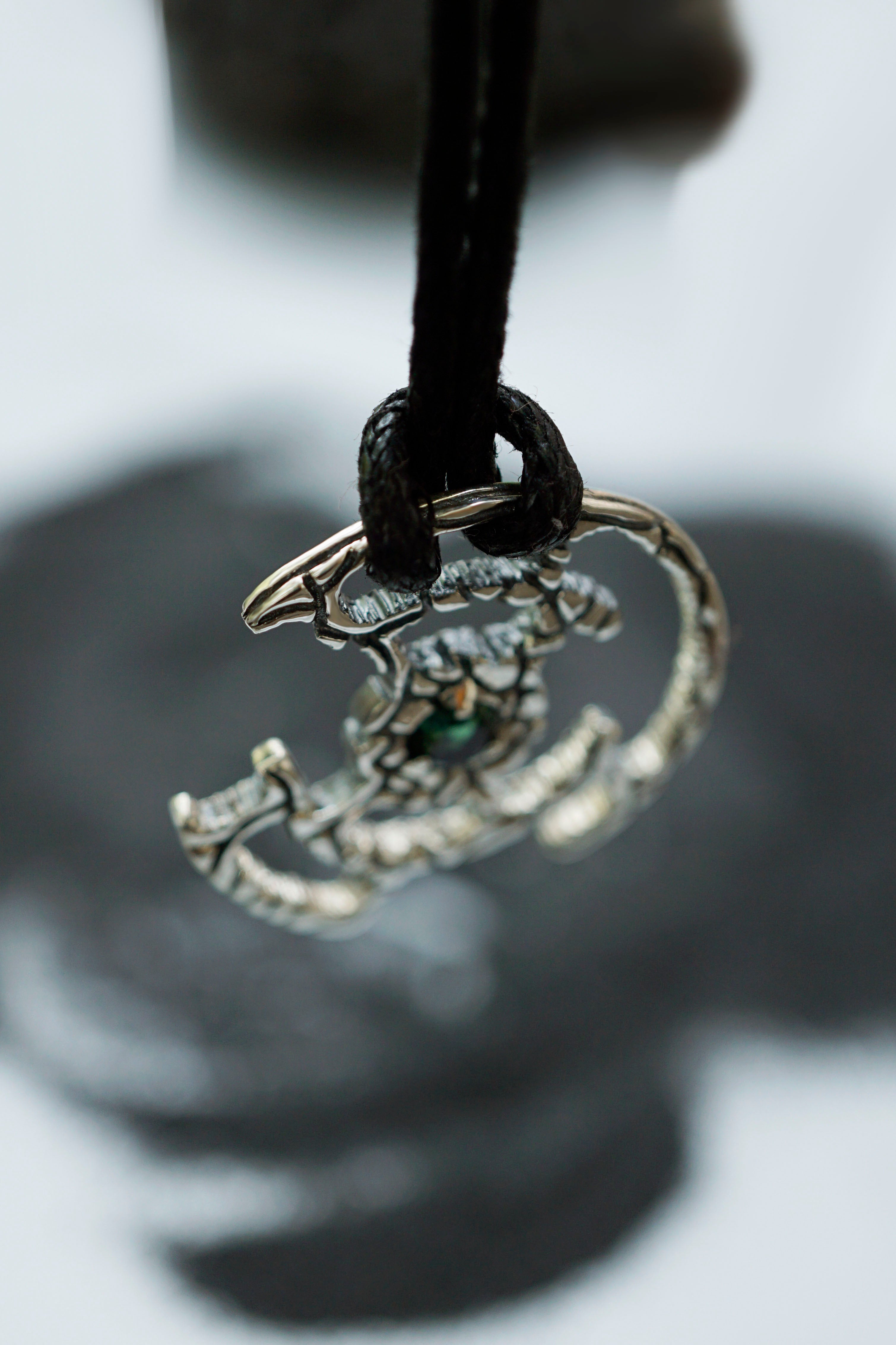 An oxidized solid sterling silver pendant with a green Tourmaline gemstone in the center. The pendant is in the shape of a circle with intricate designs. The pendant is hanging from a black cord. Cyberpunk design jewelry by Moonique.