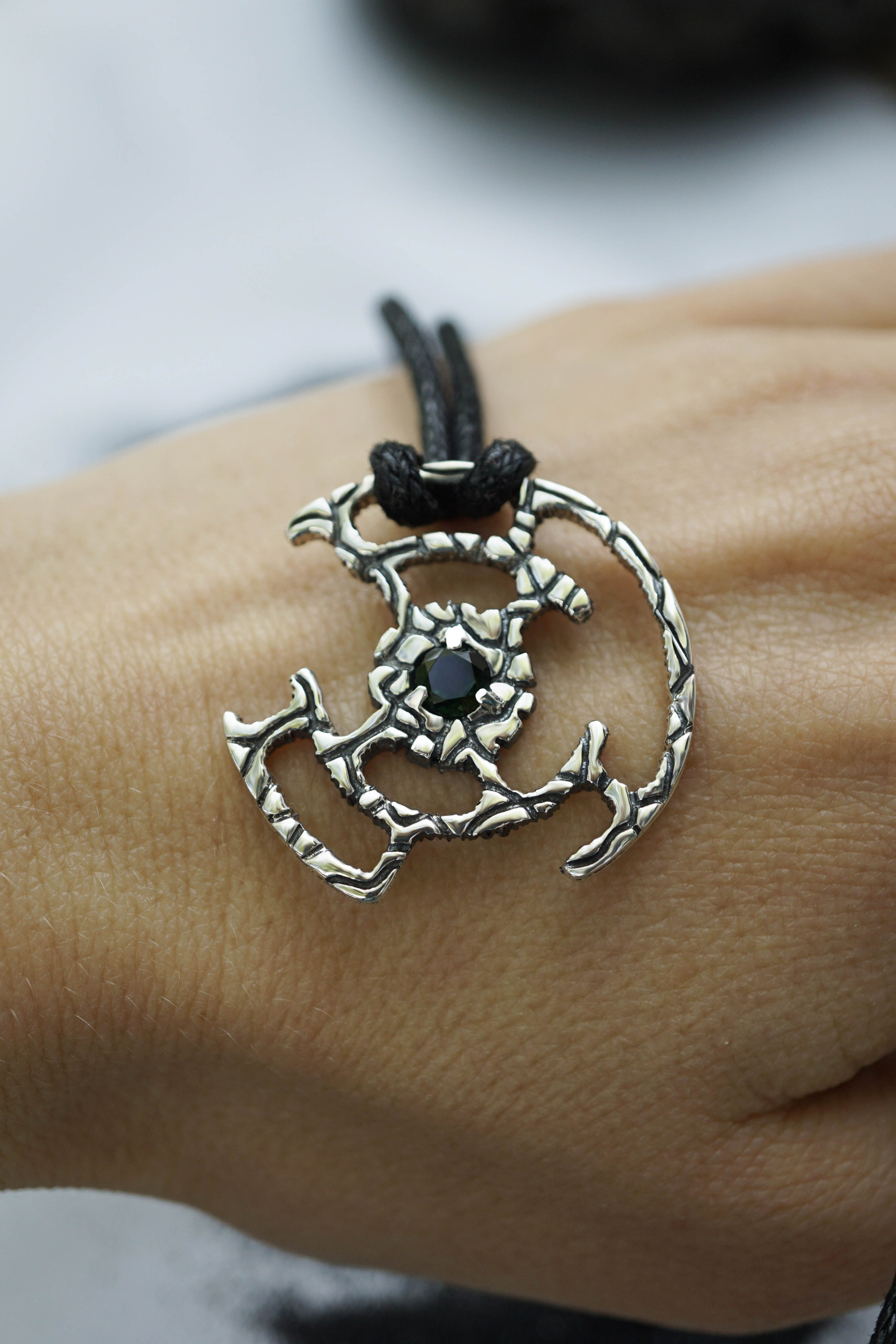 An oxidized solid sterling silver pendant with a green Tourmaline gemstone in the center. The pendant is in the shape of a circle with intricate designs. The pendant is hanging from a black cord. Cyberpunk design jewelry by Moonique.